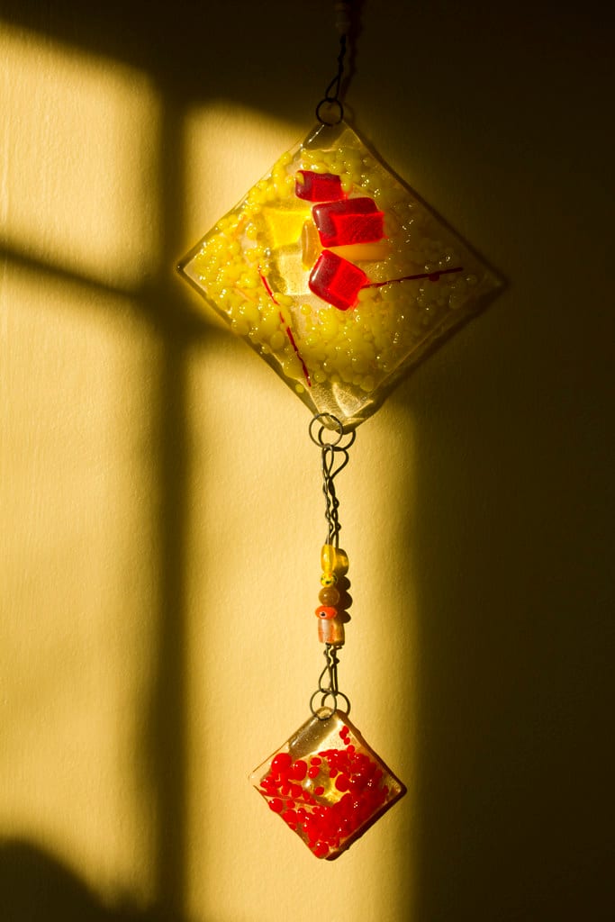 Two diamond-shaped fused glass pendant pieces hanging, one yellow with red frit details, one red, made with kiln-formed glass techniques