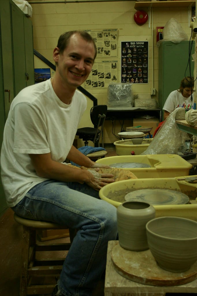 A row of pottery wheels in a busy studio class, a student throwing at the nearest one