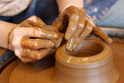 A potter sitting at a pottery wheel, hands on clay, from a slight angle showing the wheel, splash pan, and the potter's full posture